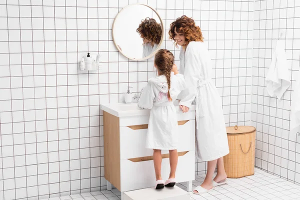 Curly woman in bathrobe looking at child near sink in modern bathroom — Stock Photo