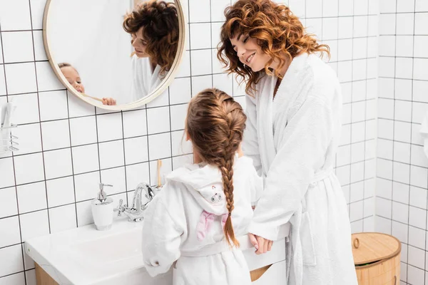 Smiling mother looking at child brushing teeth near sink and mirror — Stock Photo