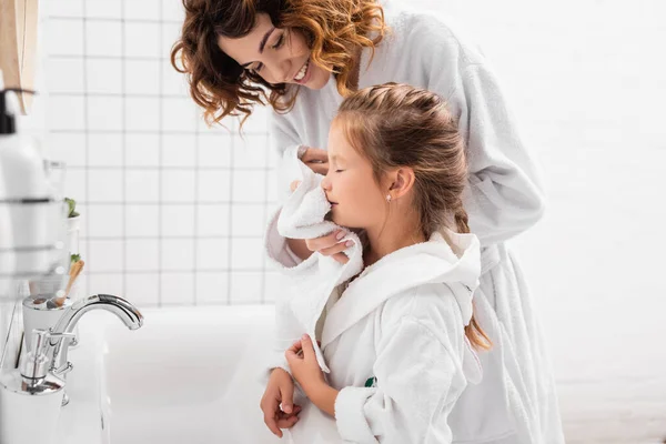 Positive woman drying face of daughter with white towel in bathroom — Stock Photo