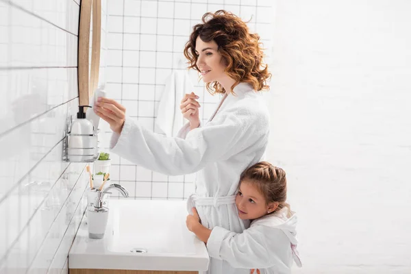 Улыбающаяся женщина берет зубную пасту рядом с ребенком в ванной комнате — стоковое фото