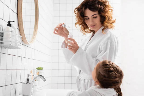 Mujer en albornoz sosteniendo suero cosmético cerca del niño en el baño — Stock Photo