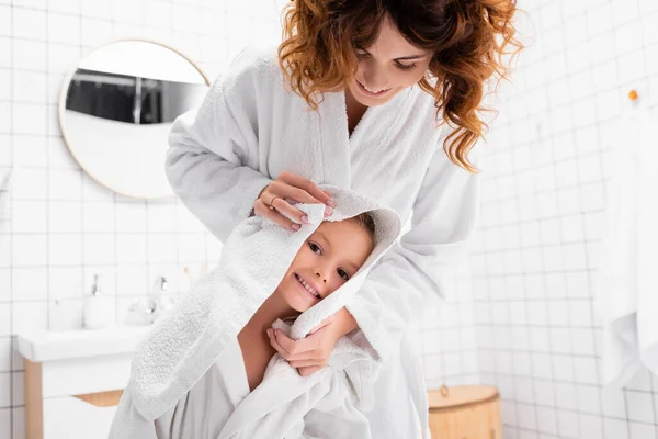 Femme souriante en peignoir séchant les cheveux de la fille avec serviette blanche dans la salle de bain — Photo de stock