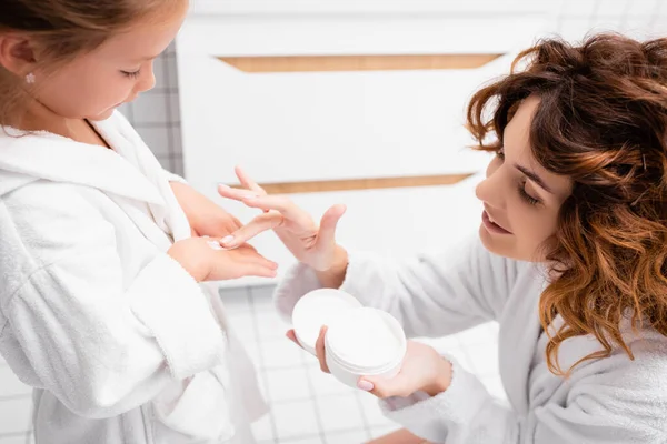 Улыбающаяся женщина наносит косметический крем на руку дочери в ванной комнате — стоковое фото