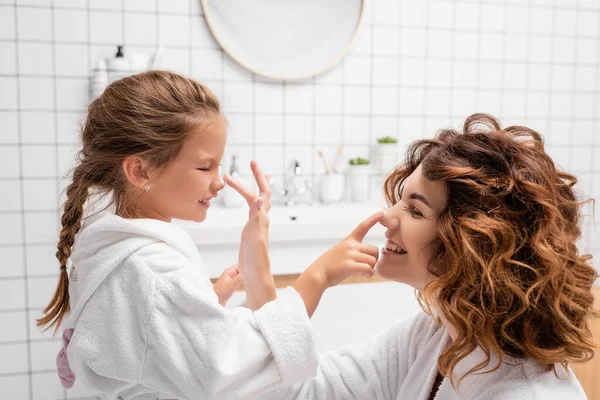 Веселая мать и ребенок наносят косметический крем на носы в ванной комнате — стоковое фото