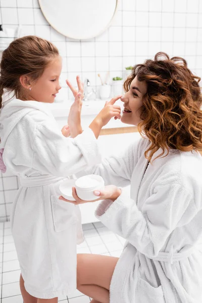 Chica sonriente aplicando crema cosmética en la nariz de la madre en el baño - foto de stock