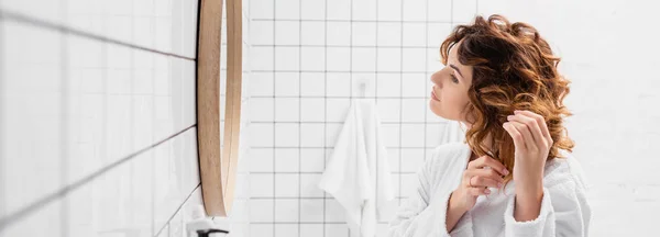 Curly woman adjusting hair near mirror in bathroom, banner — Stock Photo