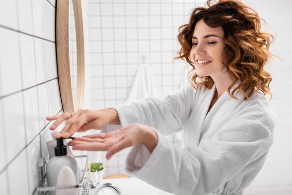 Mulher alegre aplicando creme cosmético em primeiro plano desfocado no banheiro — Fotografia de Stock
