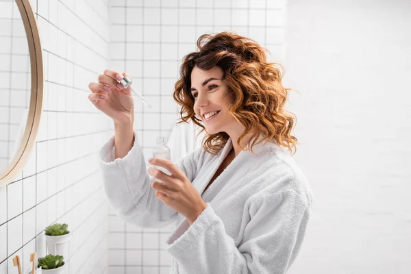 Усміхнена жінка в халаті тримає косметичну сироватку у ванній — стокове фото