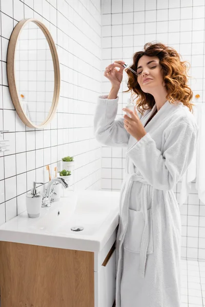 Улыбающаяся женщина в халате наносит косметическую сыворотку в ванной комнате — стоковое фото