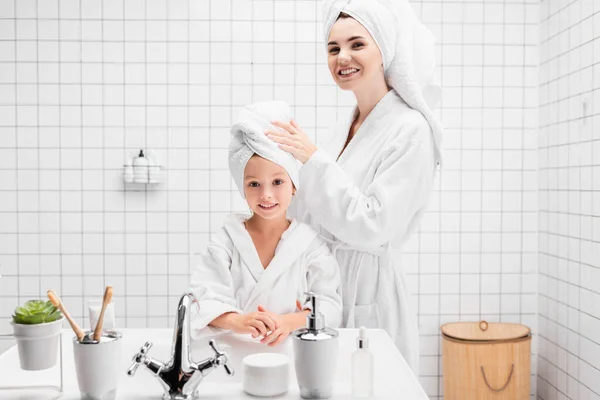 Fröhliche Mutter und Kind mit Handtüchern auf dem Kopf, die im Badezimmer in die Kamera schauen — Stockfoto