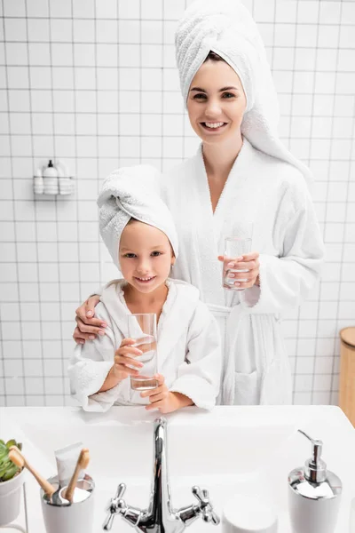 Sonrientes madre e hija en albornoces y toallas sosteniendo vasos de agua en el baño - foto de stock