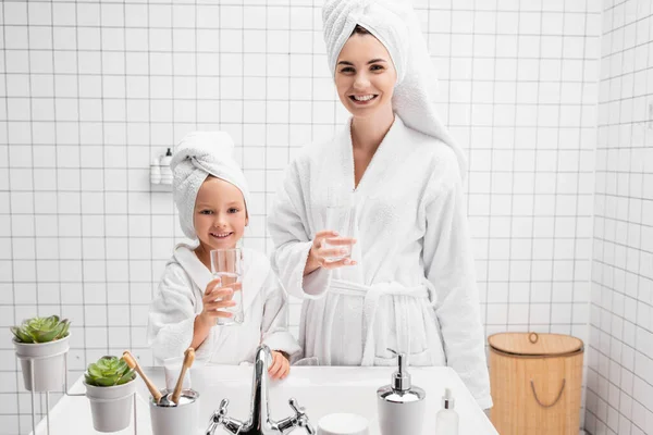 Позитивные мать и дочь держат стаканы воды в ванной комнате — стоковое фото