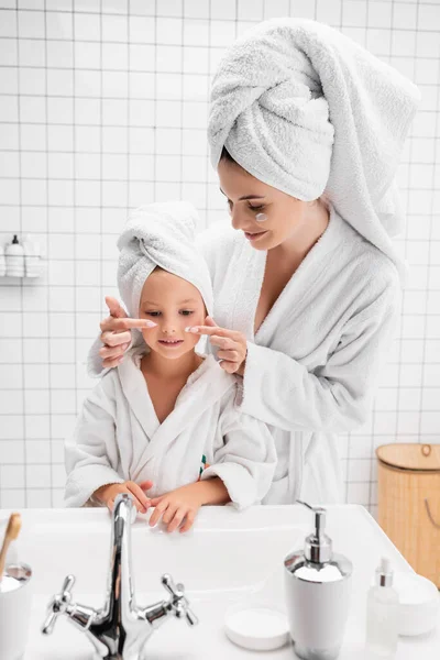 Madre aplicando crema facial en las mejillas de la hija en el baño - foto de stock