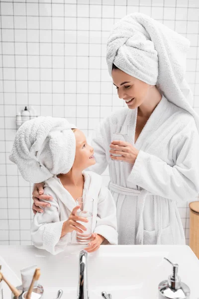 Madre sonriente en albornoz y toalla sosteniendo vaso de agua cerca de la hija en el baño - foto de stock