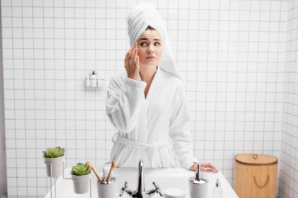 Mujer en albornoz y toalla con la mano cerca de la cara en el baño - foto de stock