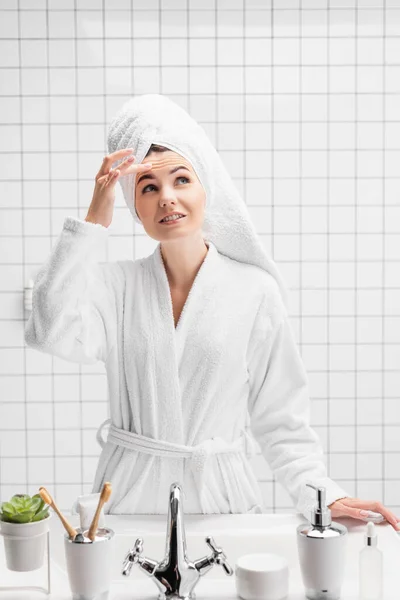Усміхнена жінка в рушнику торкається лоба у ванній — стокове фото