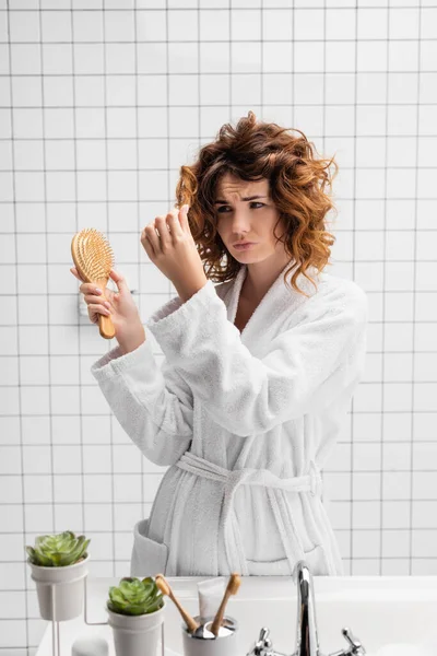 Upset woman holding hair brush and looking at hair in bathroom — Stock Photo