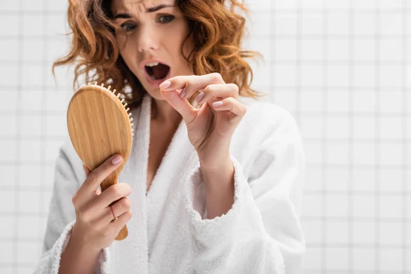 Brosse à cheveux à la main de femme étonnée sur fond flou dans la salle de bain sur fond flou — Photo de stock