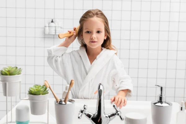 Girl in bathrobe brushing hair near sink in modern bathroom — Stock Photo