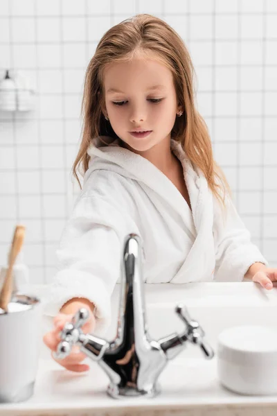 Fille ouvrant l'eau près de l'évier sur le premier plan flou dans la salle de bain — Photo de stock