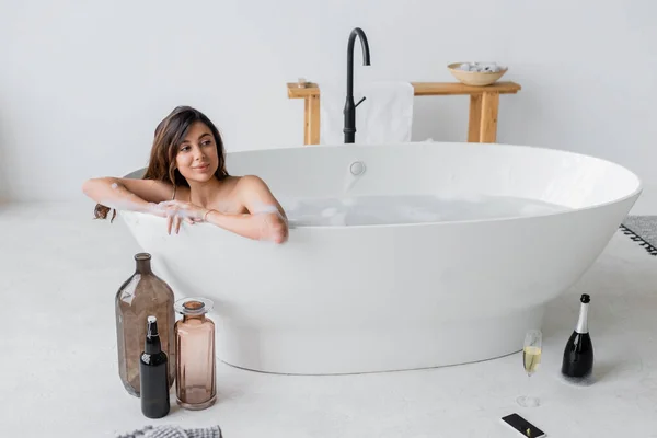 Femme souriante assise dans la baignoire près du champagne et smartphone sur le sol — Photo de stock