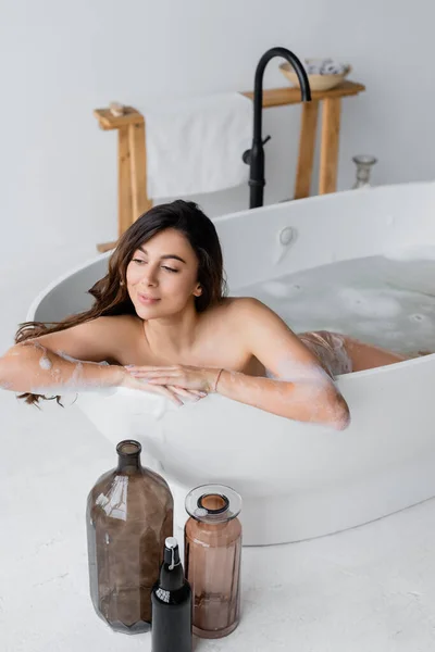 Mulher morena relaxante na banheira perto de garrafas decorativas — Fotografia de Stock
