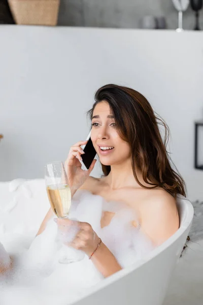Улыбающаяся женщина в пене разговаривает по смартфону и держит бокал шампанского в ванной — стоковое фото