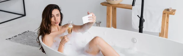 Жінка з келихом шампанського бере селфі зі смартфоном у ванній, банер — стокове фото