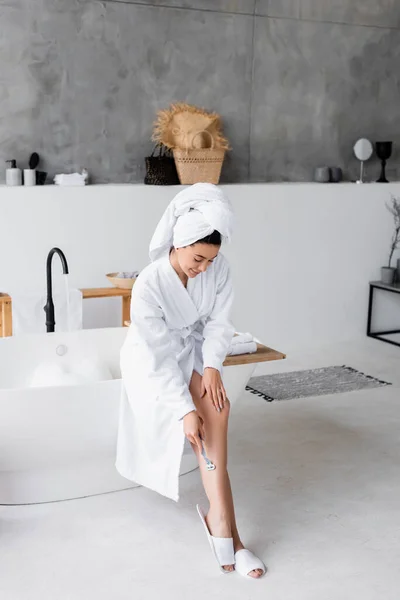 Young woman shaving leg near bathtub at home — Stock Photo