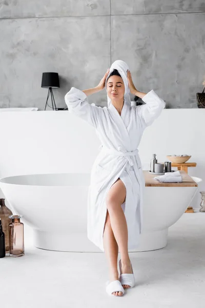 Giovane donna in accappatoio bianco e asciugamano seduta ad occhi chiusi sulla vasca da bagno — Foto stock