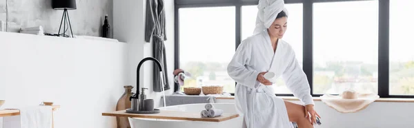 Femme en peignoir appliquant crème cosmétique sur la jambe dans la salle de bain, bannière — Photo de stock
