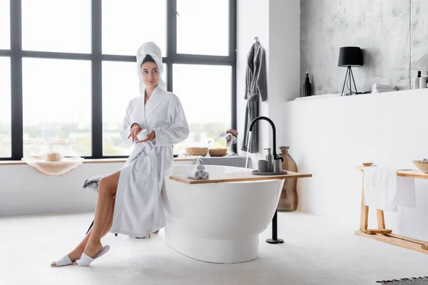 Woman in towel and bathrobe holding cosmetic cream in modern bathroom — Stock Photo