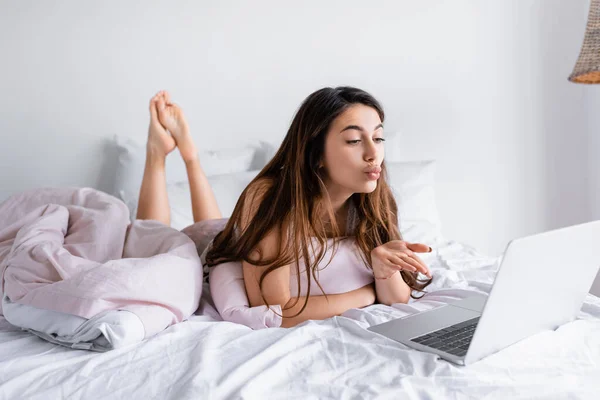 Woman blowing air kiss at laptop on blurred foreground on bed — Stock Photo