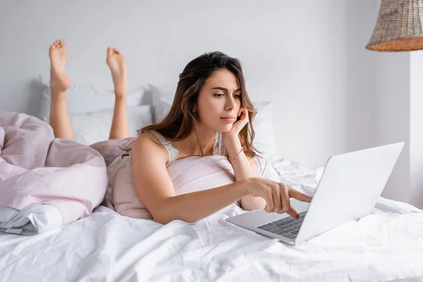 Pensive woman pointing at laptop while lying on bed — Stock Photo