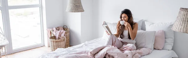 Mujer joven con taza de libro de lectura de café en la cama, pancarta - foto de stock