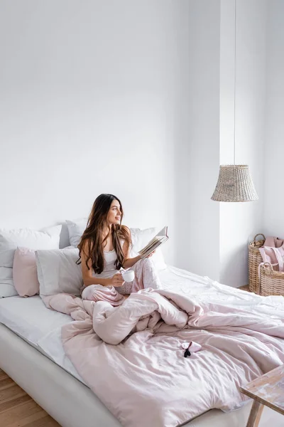 Smiling woman with coffee cup holding book on bed — Stock Photo