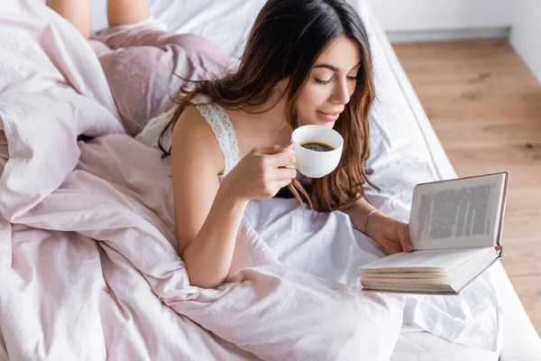 Donna in pigiama in possesso di una tazza di caffè e libro di lettura sul letto — Foto stock