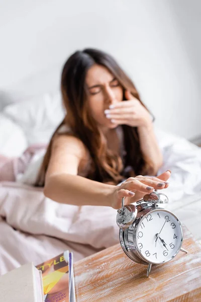 Schläfrige Frau auf verschwommenem Hintergrund schaltet Wecker auf Nachttisch aus — Stockfoto