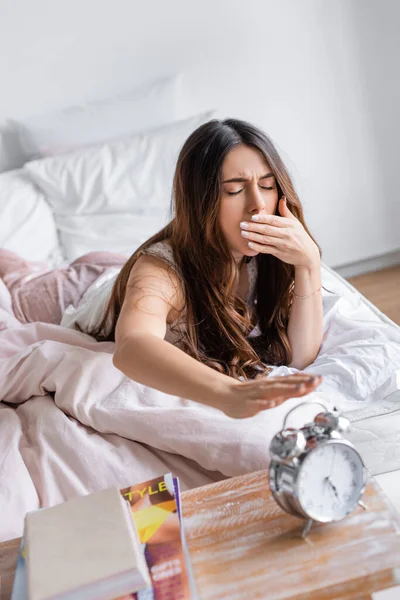 Yawning woman pulling hand to alarm clock on blurred foreground in bedroom — Stock Photo