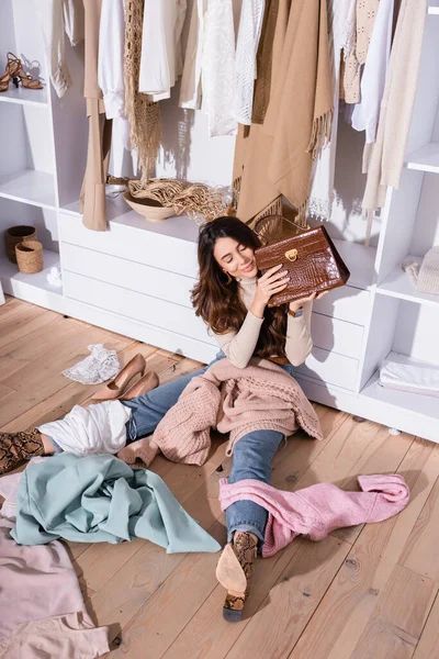 Visão de alto ângulo de mulher sorridente segurando bolsa perto de roupas no chão no guarda-roupa — Fotografia de Stock