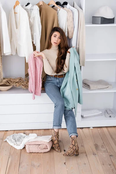 Woman looking at camera while holding clothes near shelves in wardrobe — Stock Photo