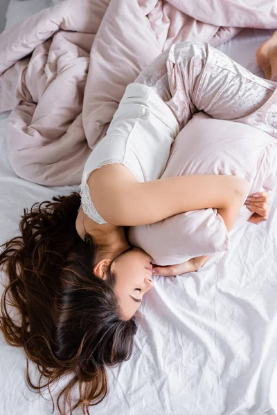 Vista superior de la mujer morena en pijama abrazando almohada mientras está acostado en la cama - foto de stock