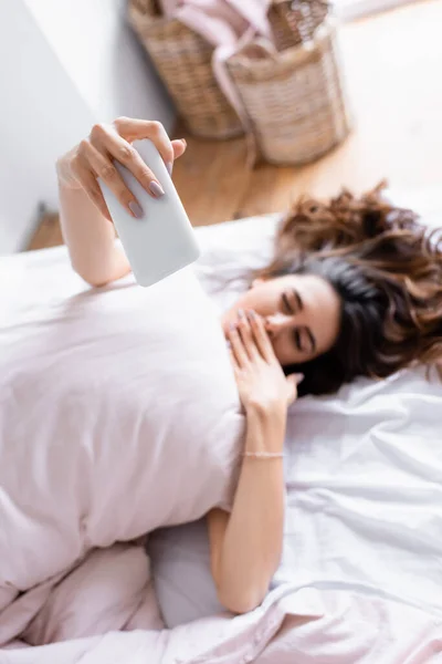 Smartphone in hand of woman taking selfie on blurred background on bed — Stock Photo