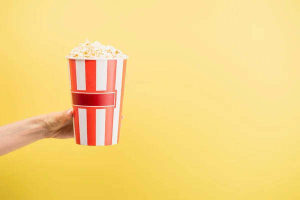 Vista parcial de la mujer sosteniendo cubo de deliciosas palomitas de maíz aislado en amarillo, concepto de cine - foto de stock