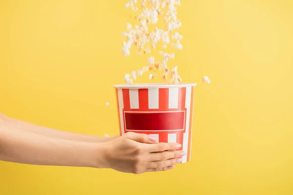 Tasty popcorn falling into striped bucket in female hands isolated on yellow, cinema concept — Stock Photo