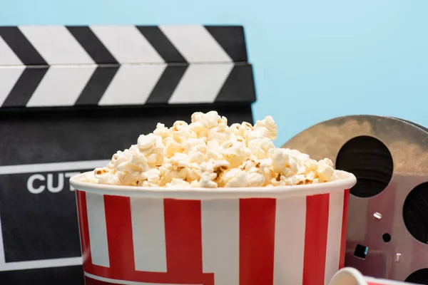Close up view of bucket with popcorn near clapperboard and film reel isolated on blue, cinema concept — Stock Photo