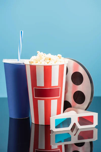 Bucket with popcorn, cup with soda, film bobbin and 3d cinema glasses on glossy surface isolated on blue — Stock Photo