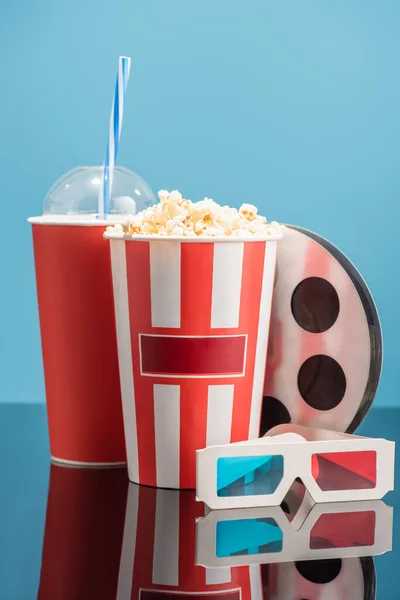 Cup of soda, bucket with popcorn, 3d glasses and film bobbin on glossy surface isolated on blue, cinema concept — Stock Photo