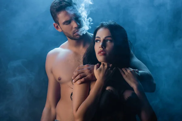 Sexy man smoking and hugging girlfriend in bra on black background with smoke — Stock Photo