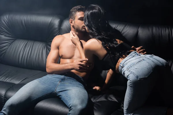 Sexy woman kissing boyfriend on jeans on couch on black background with smoke — Stock Photo